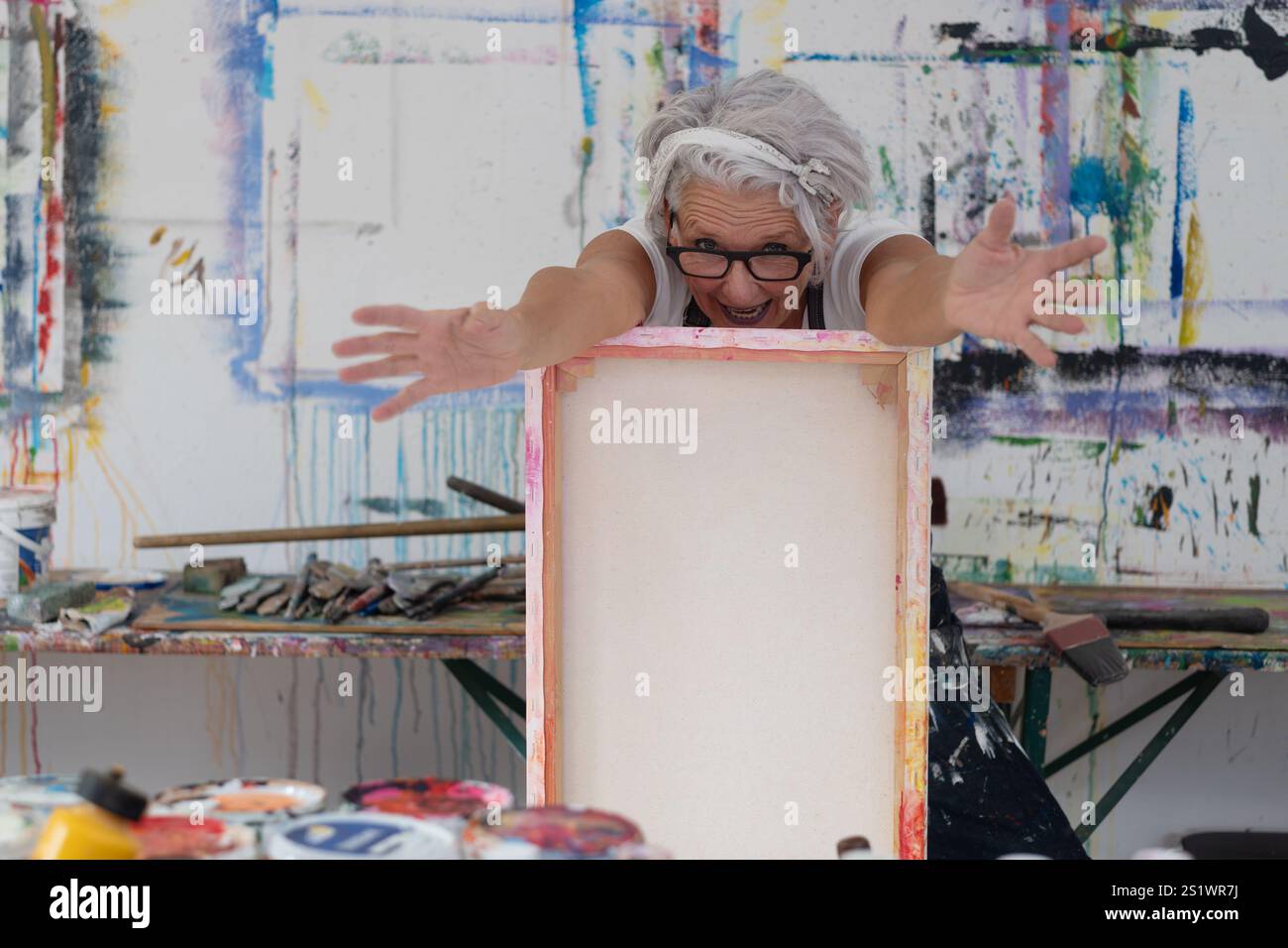 Artista donna anziana e matta, espressiva, che trasuda energia e creatività, si appoggia su una tela colorata nel suo studio d'arte. Il suo gioioso entusiasmo radia Foto Stock
