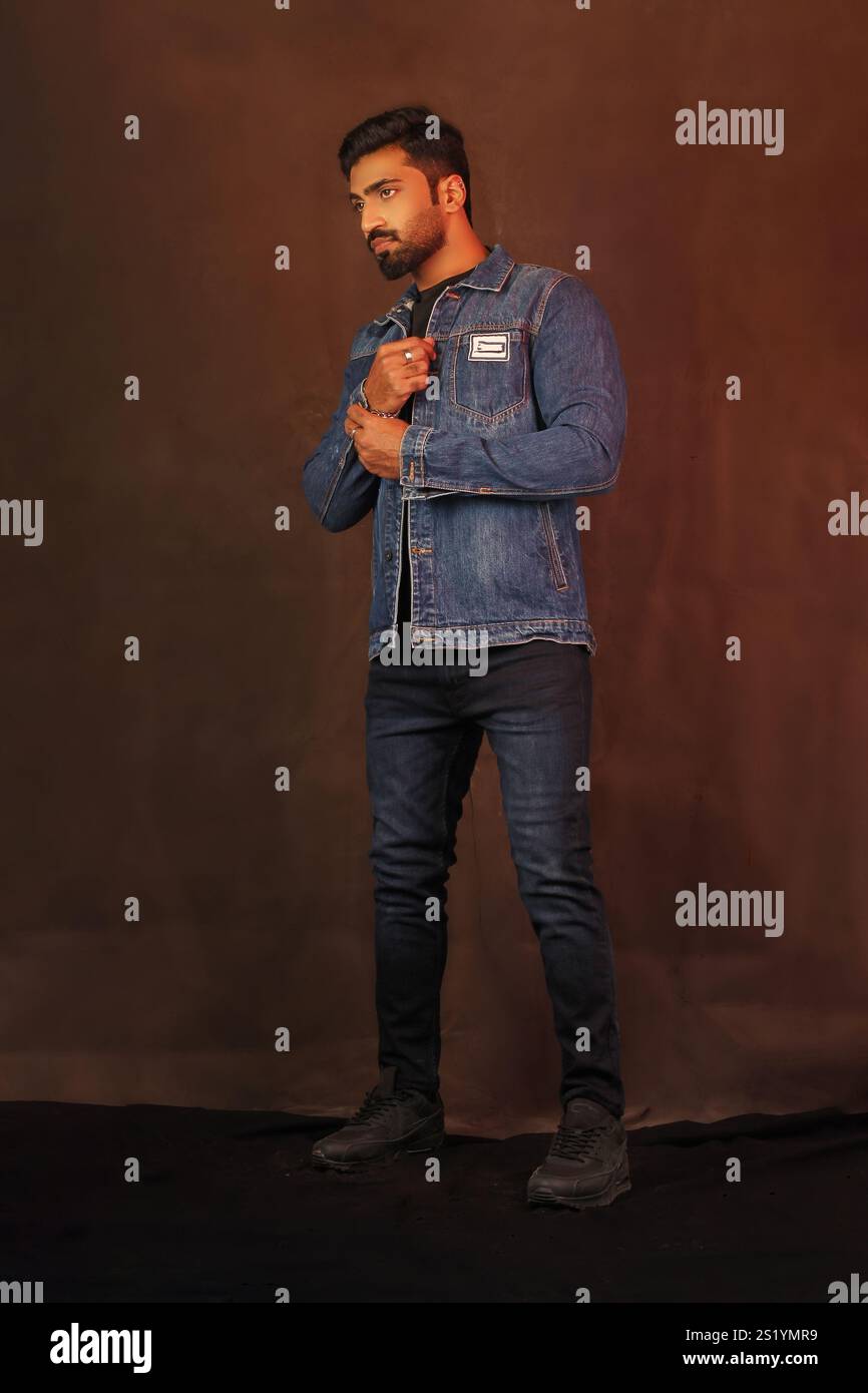 Un modello indiano maschile in posa con giacca in denim e jeans su sfondo grigio a figura intera Foto Stock