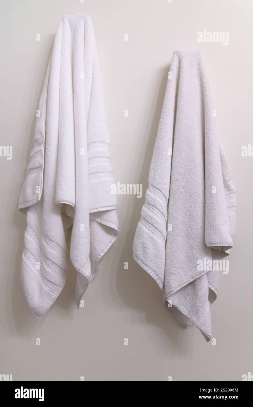 Due asciugamani da bagno bianchi appesi su ganci su una parete bianca Foto Stock