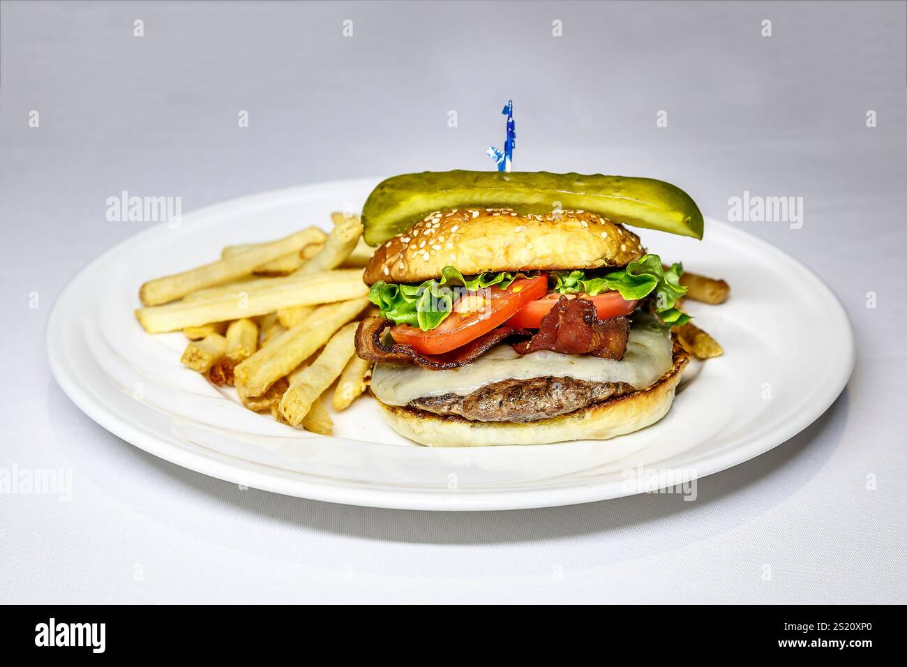 Un hamburger con una lancia di sottaceti sopra. L'hamburger viene servito su un piatto bianco con patatine fritte a lato Foto Stock