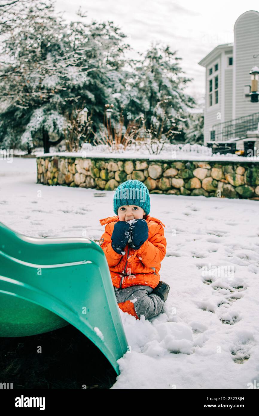 Bambino con giacca arancione e cappello verde seduto sulla neve Foto Stock