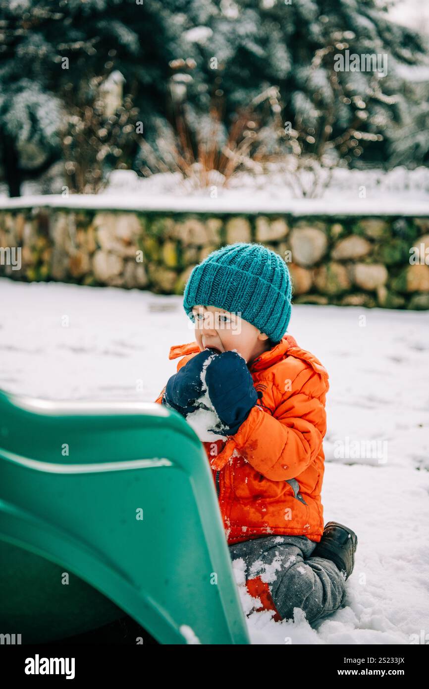Bambino che mangia la neve vicino allo scivolo verde Foto Stock