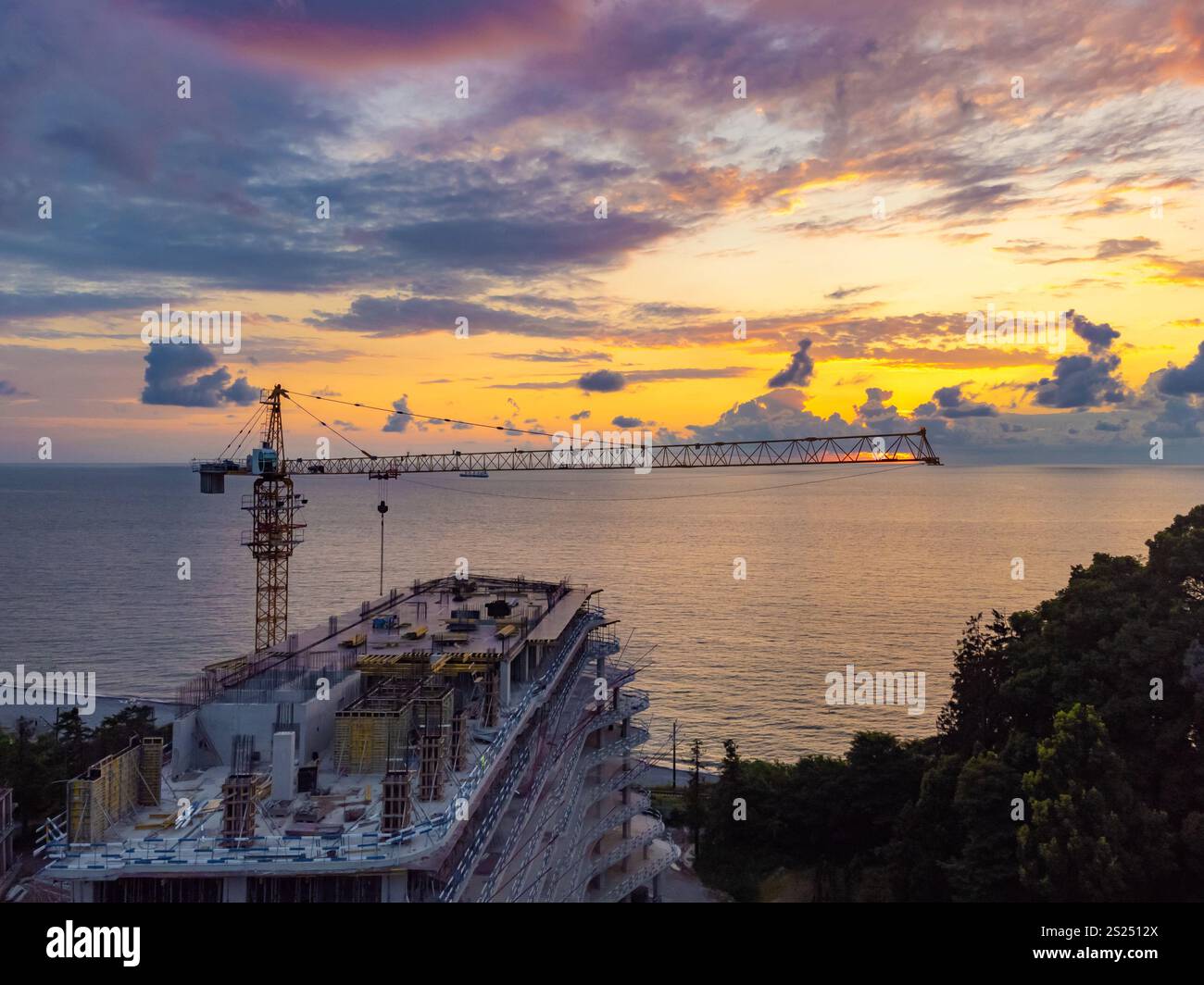 Vista droni di un edificio a più piani in costruzione e di una gru da costruzione sullo sfondo di un bellissimo tramonto sul mare. Concetto di costruzione Foto Stock