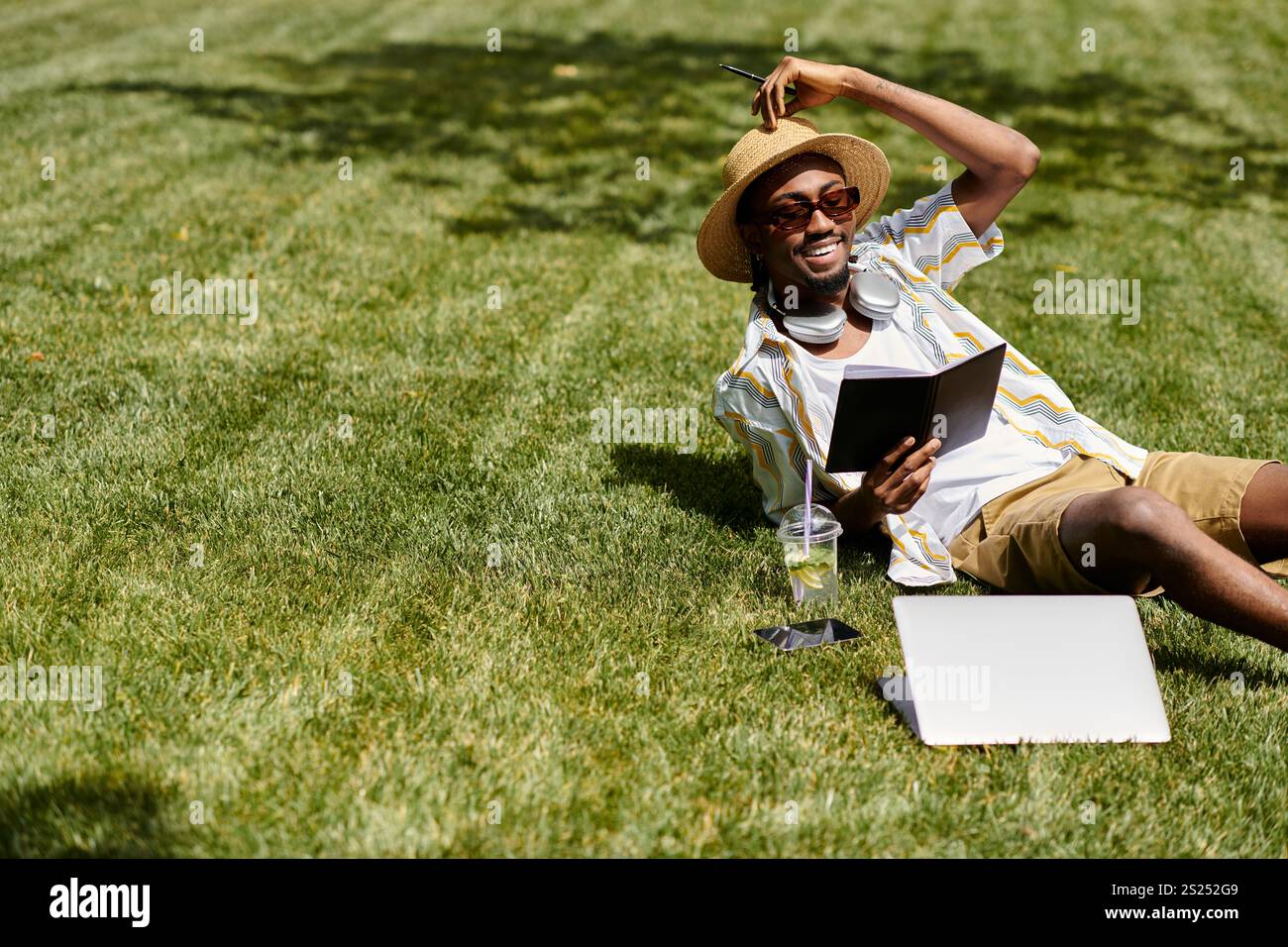 Un bel giovane si rilassa sull'erba, lavorando al suo portatile con un drink in mano. Foto Stock