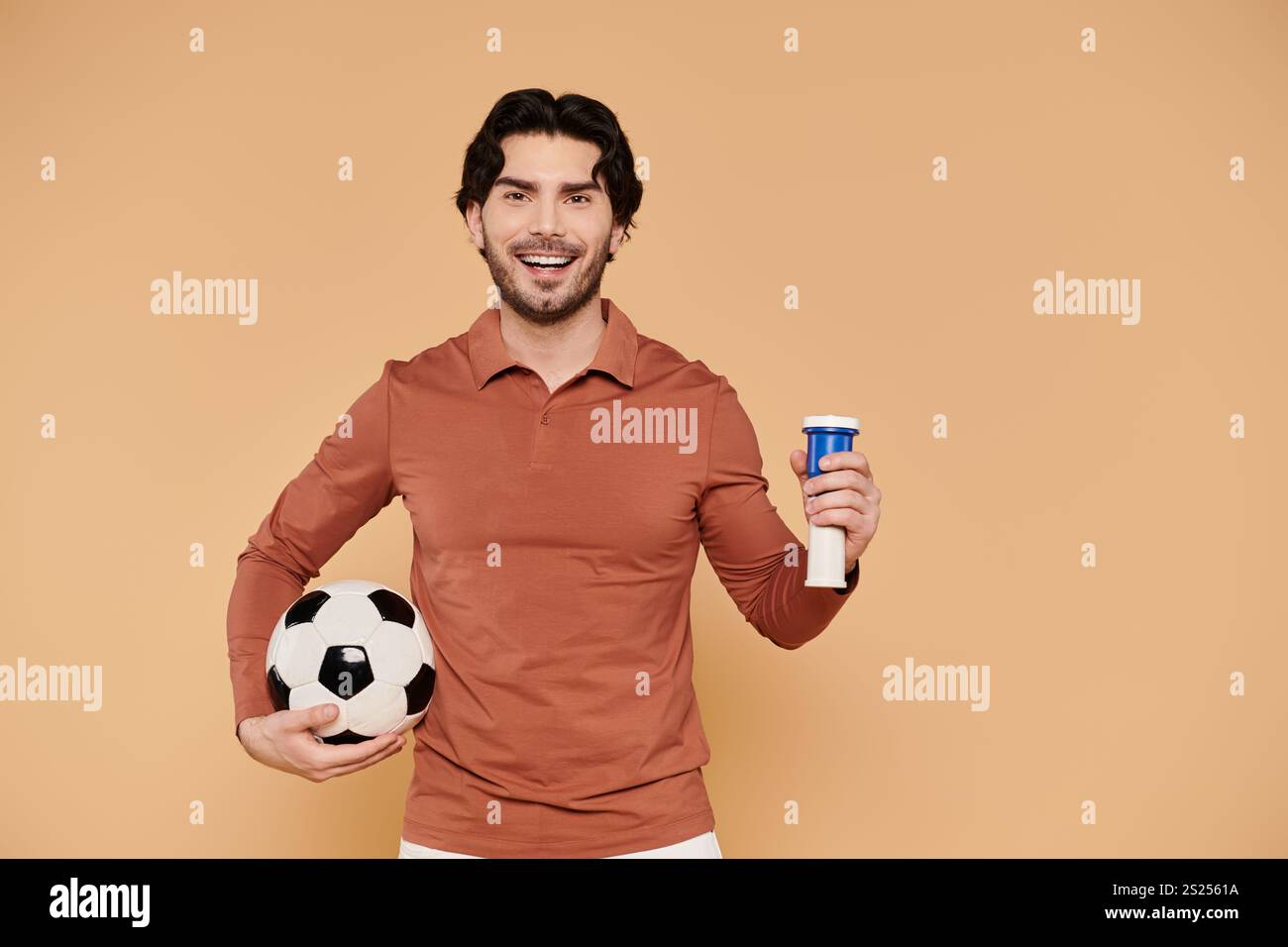 Un giovane gioioso sorride tenendo in mano una palla da calcio e un drink nell'altra. Foto Stock