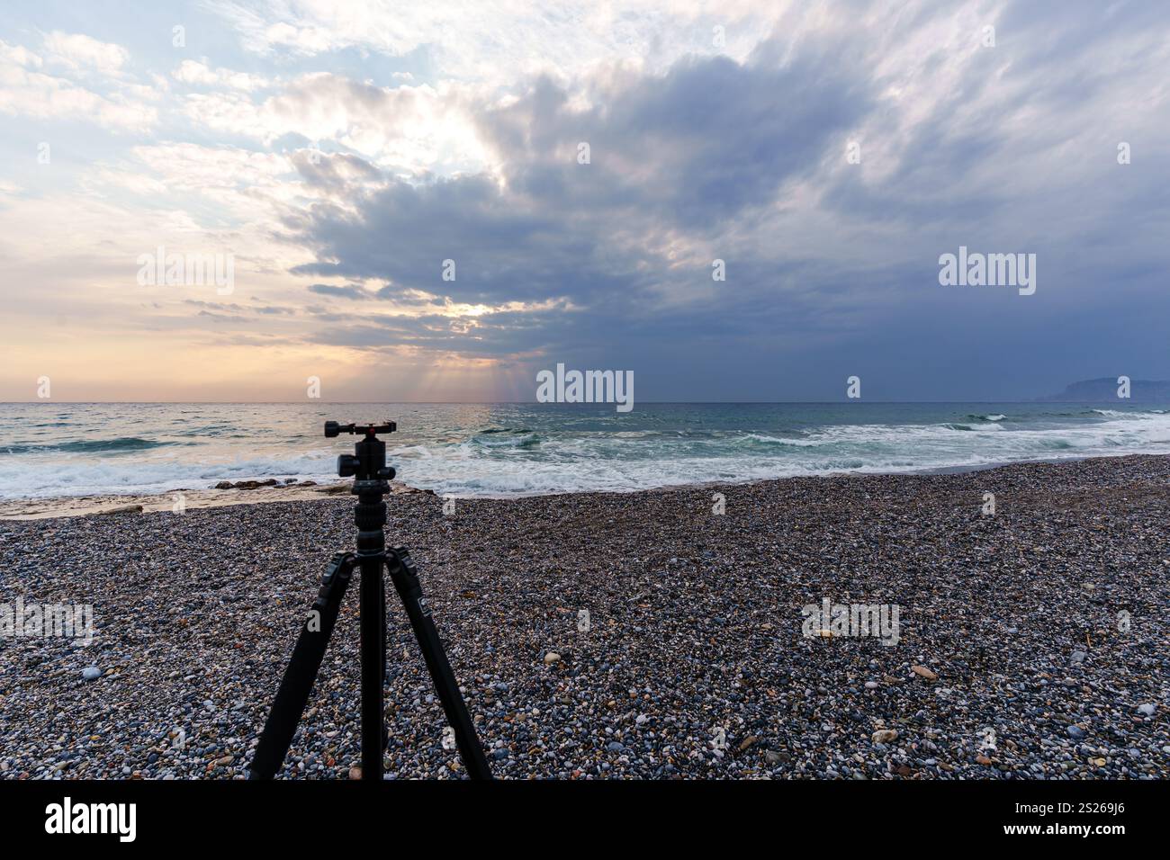 Una tranquilla spiaggia all'alba con un treppiede per fotocamera su una riva di ciottoli, che cattura le onde tranquille e il cielo esteso. Ideale per i concetti di fotografia Foto Stock