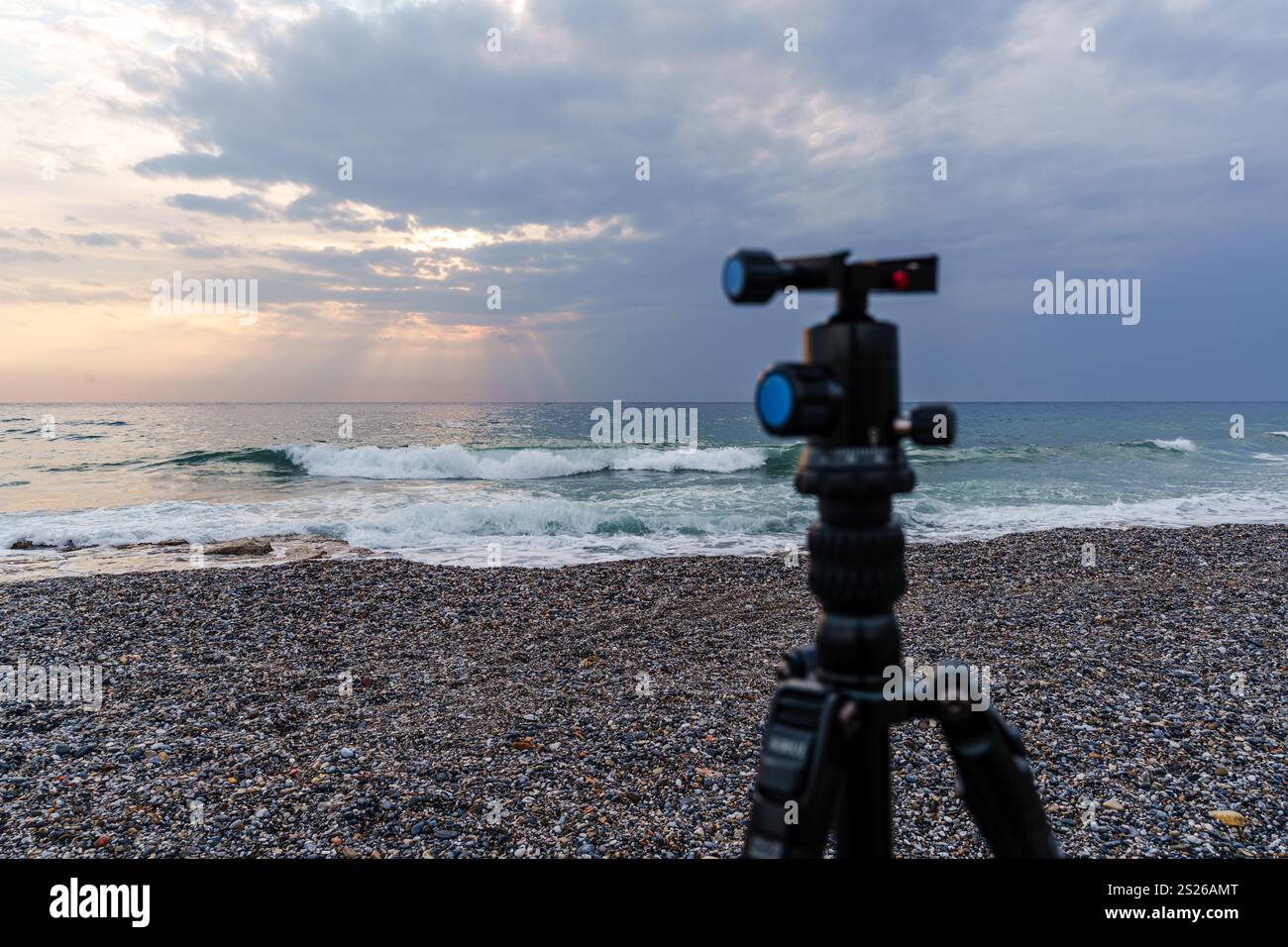 Una tranquilla spiaggia all'alba con un treppiede per fotocamera su una riva di ciottoli, che cattura le onde tranquille e il cielo esteso. Ideale per i concetti di fotografia Foto Stock
