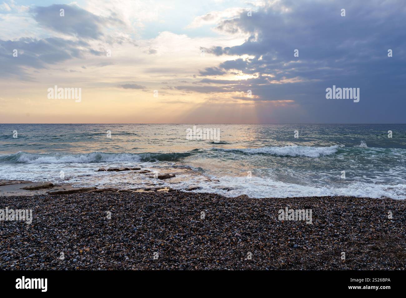 Tranquillo scenario di una spiaggia di ciottoli al tramonto. Il cielo è pieno di nuvole spettacolari e raggi del sole si estendono attraverso l'oceano, creando un clima tranquillo e rilassante Foto Stock