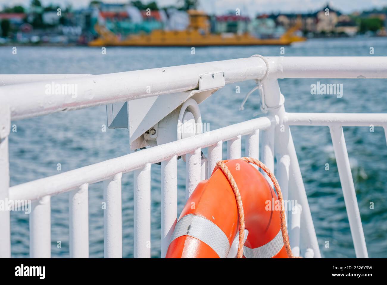 Salvagente arancione con strisce riflettenti appese su ringhiera bianca del traghetto che naviga su acque blu con paesaggio urbano e traghetto giallo in sfocatura Foto Stock