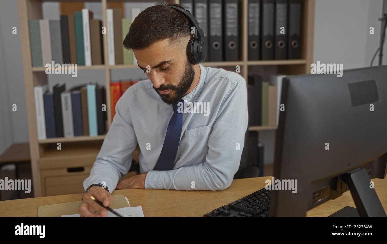 Uomo ispanico che indossa le cuffie e scrive alla sua scrivania in un ufficio pieno di libri e file. Foto Stock
