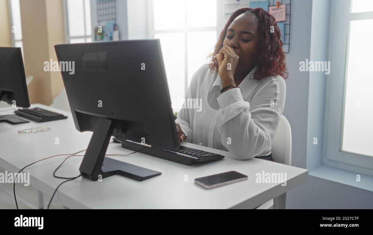 Donna afroamericana che lavora al chiuso in una stanza d'ufficio, guardando un computer mentre guarda premurosa. Foto Stock