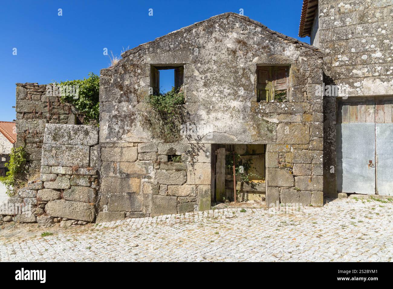 Facciata anteriore di una vecchia casa di granito abbandonata con pantaloni che crescono fuori dalla finestra, manca la porta d'ingresso, cielo blu. Foto Stock