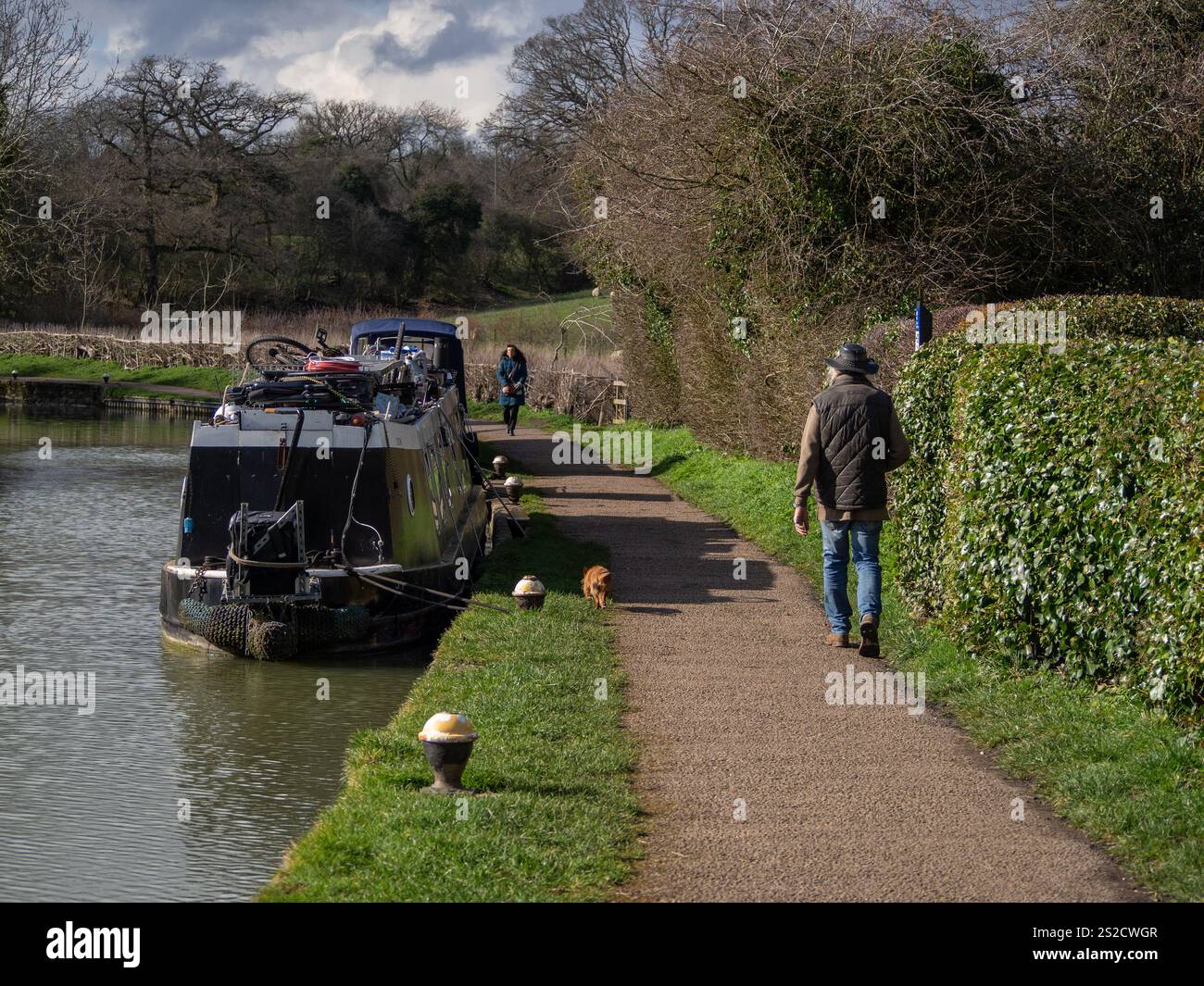 Passeggia per cani su un sentiero lungo il canale, passando davanti a una barca ormeggiata in una soleggiata giornata invernale, Stoke Bruene, Northamptonshire, Regno Unito Foto Stock