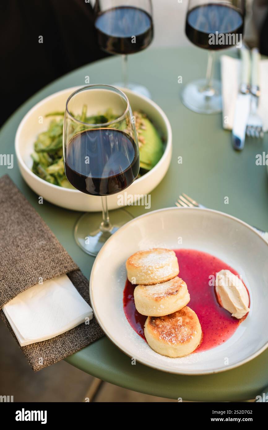 Pancake di Quark (Syrniki) con marmellata di fragole, un'insalata di avocado e tre bicchieri di vino rosso su un tavolo in un caffè all'aperto in estate Foto Stock