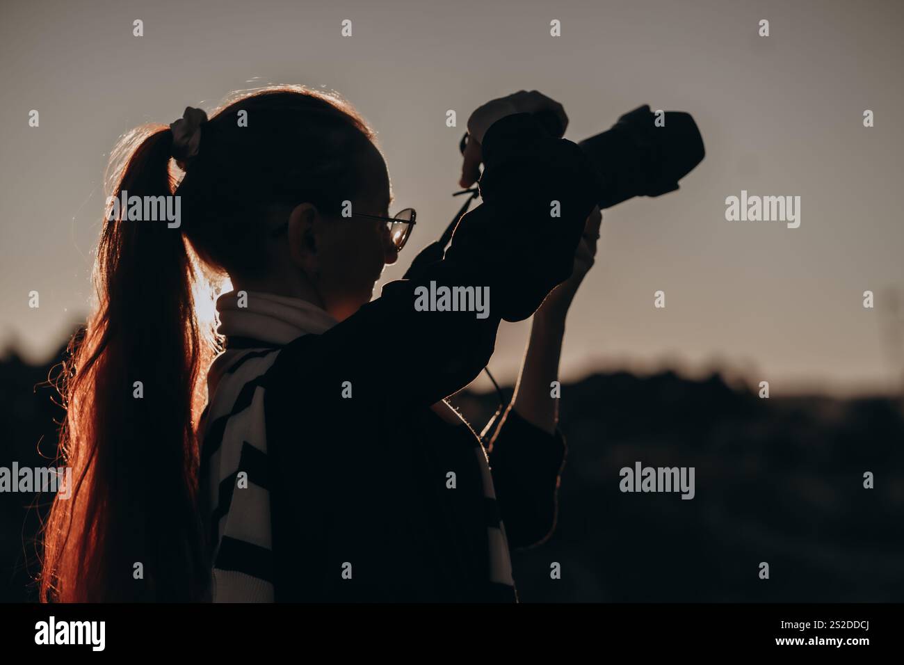 La silhouette di una fotografa che tiene in mano con sicurezza una fotocamera sulla spalla si distingue sullo sfondo di un tramonto sereno. Il suo lungo pony Foto Stock