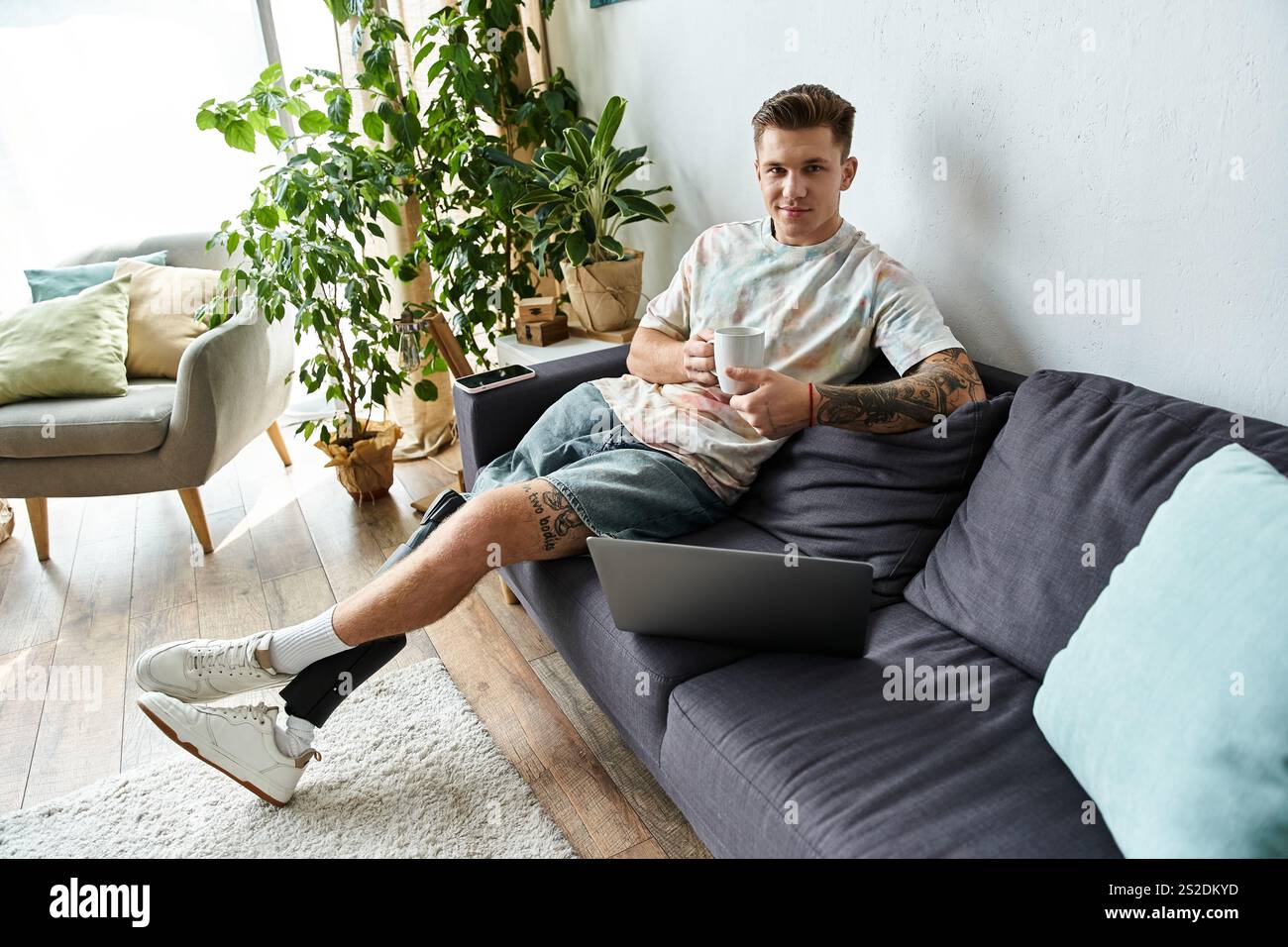 Un giovane e bello uomo si siede comodamente su un divano, gustando un caffè mentre lavora su un notebook. Foto Stock