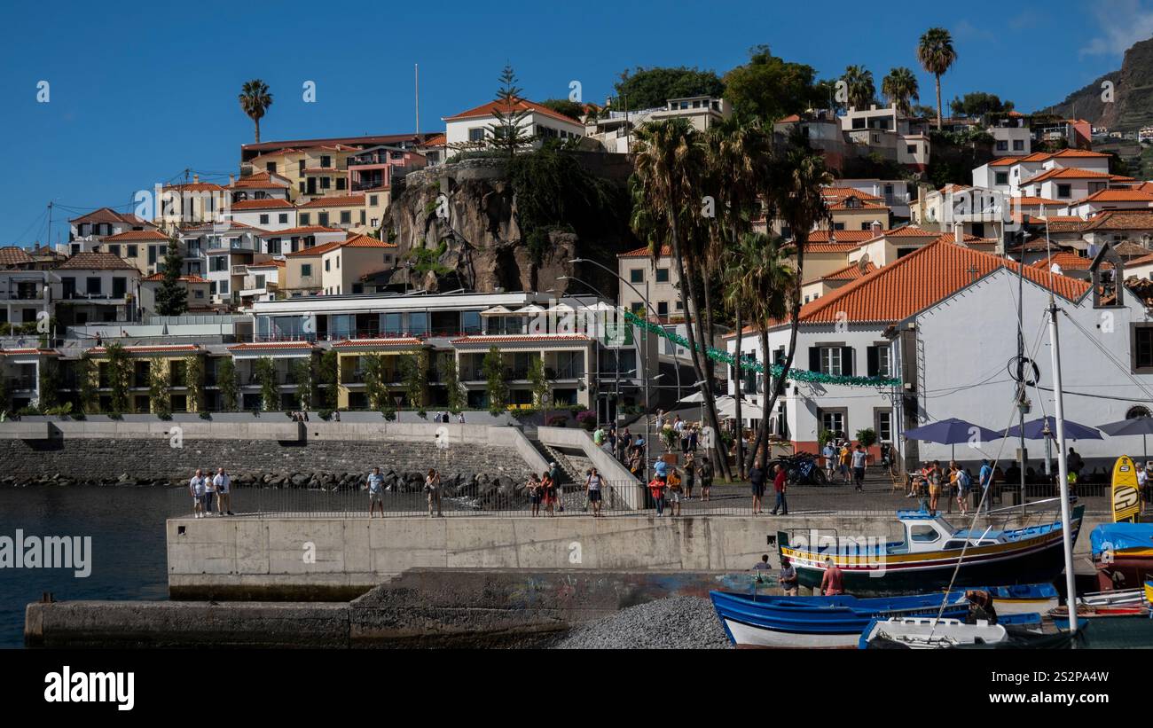 Un'affascinante cittadina costiera con case colorate su una collina, palme e un vivace porto con barche da pesca e turisti sotto un blu brillante Foto Stock