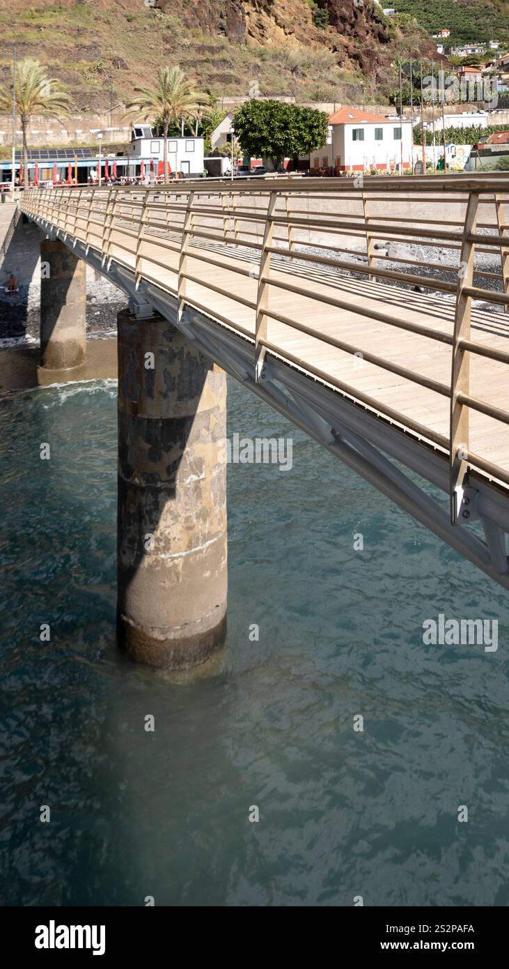 Un ponte pedonale con ringhiere in metallo sulle acque blu calme, sostenuto da pilastri di cemento, con un villaggio costiero e scogliere rocciose sullo sfondo Foto Stock