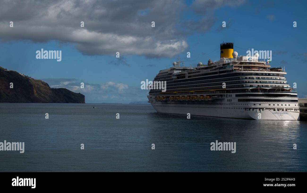Una grande nave da crociera attraccata in un porto calmo sotto un cielo parzialmente nuvoloso, con una costa rocciosa visibile sullo sfondo. Foto Stock
