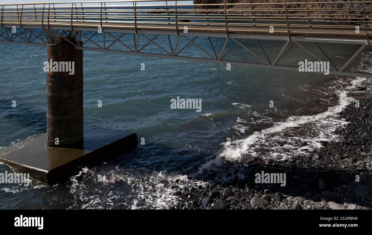 Un ponte pedonale in metallo che si estende su una costa rocciosa, con onde delicate che si infrangono sulle pietre e il mare blu sotto la luce del sole. Foto Stock