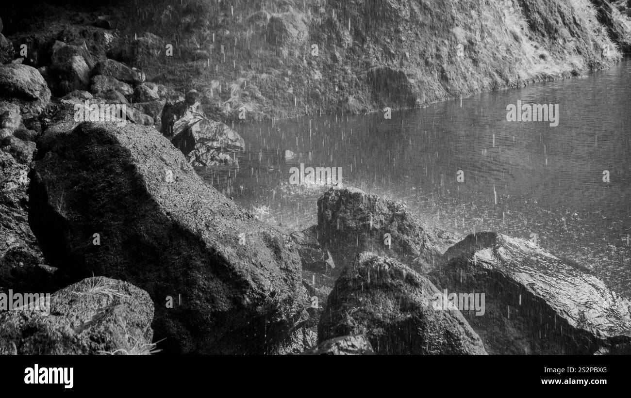 Un'immagine in bianco e nero della pioggia che cade su grandi rocce vicino a un corpo d'acqua calmo, creando increspature e migliorando la texture naturale della scena. Foto Stock