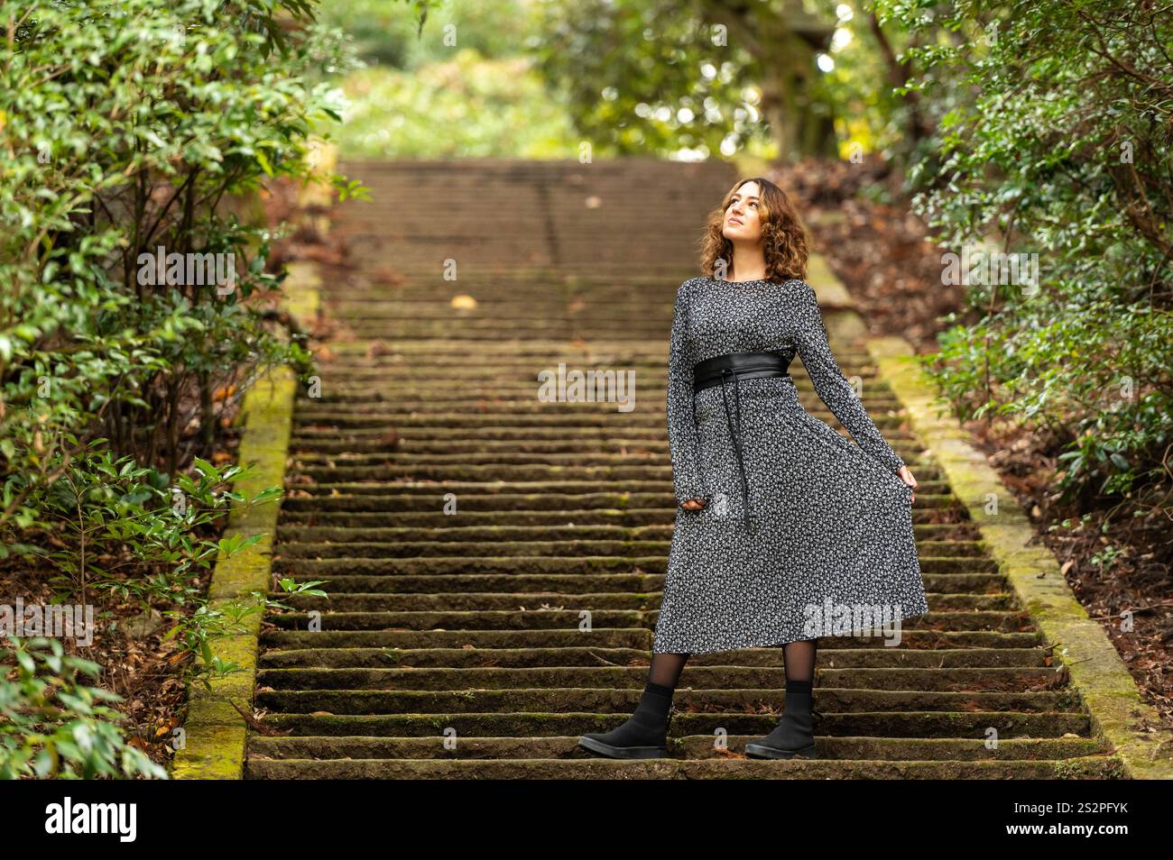 Donna che indossa un abito floreale in piedi con sicurezza su scale di pietra mossy in un lussureggiante ambiente verde della foresta. Foto Stock