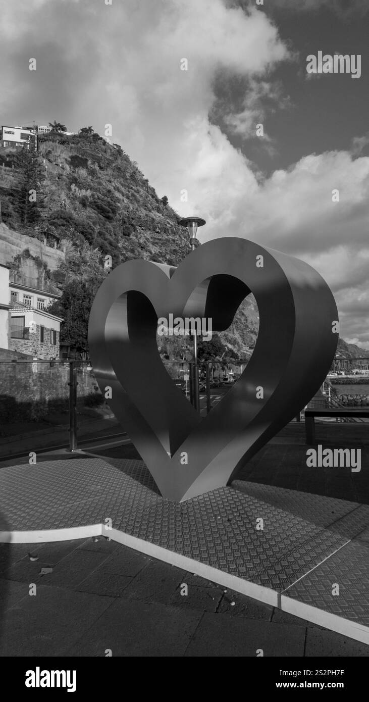 Una fotografia in bianco e nero di una grande scultura in metallo a forma di cuore in un ambiente urbano all'aperto. Lo sfondo presenta una collina con edifici Foto Stock