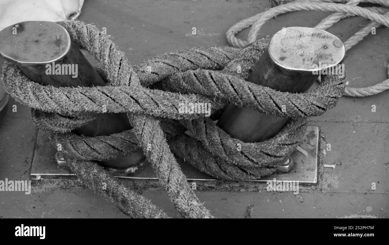 Primo piano bianco e nero di spesse corde avvolte e legate intorno a due dissuasori di metallo su un ponte della nave, sottolineando la consistenza e i dettagli nautici. Foto Stock