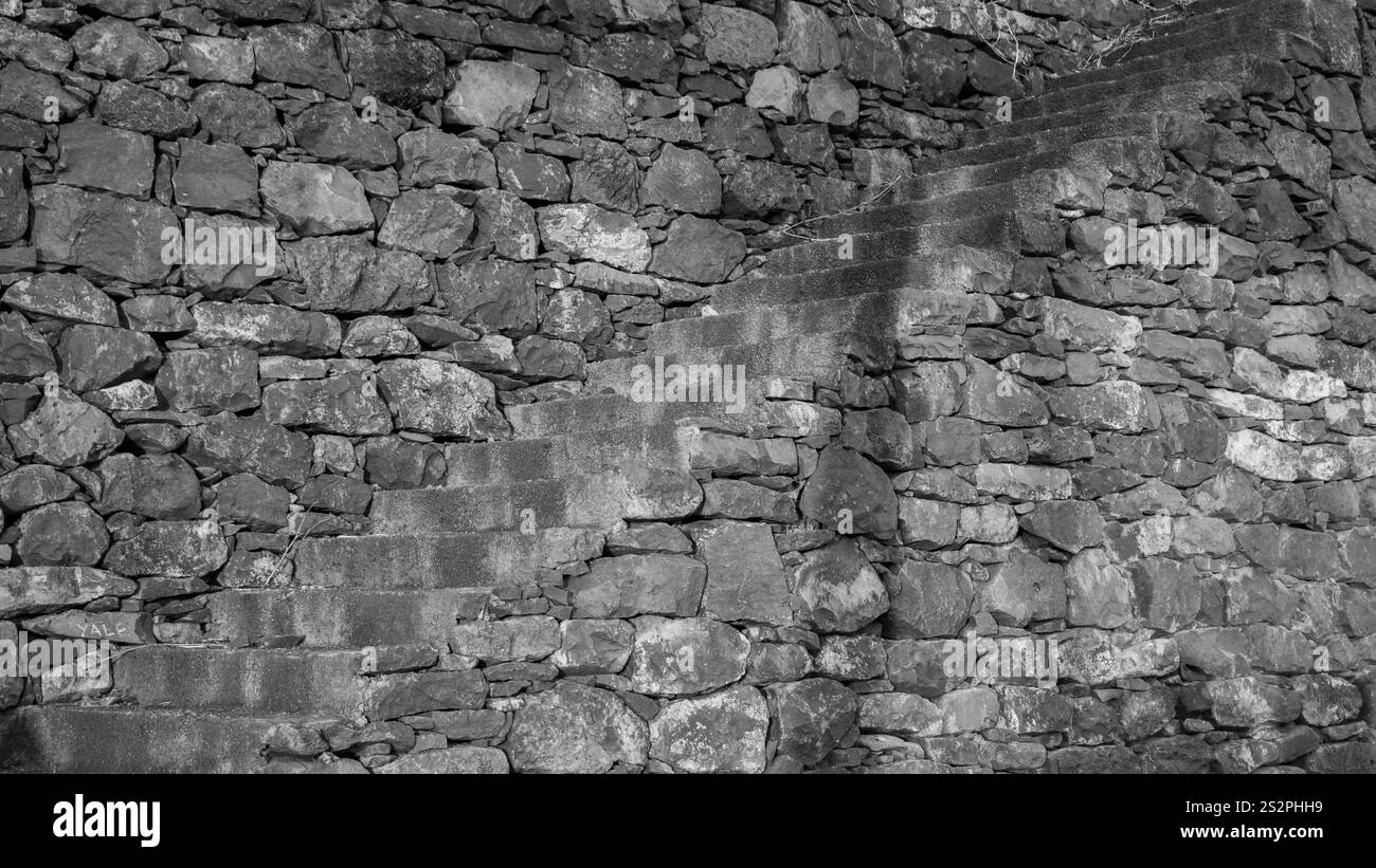 Un'immagine in bianco e nero di un muro di pietra con una scala incorporata, che mostra muratura rustica e una composizione angolare Foto Stock