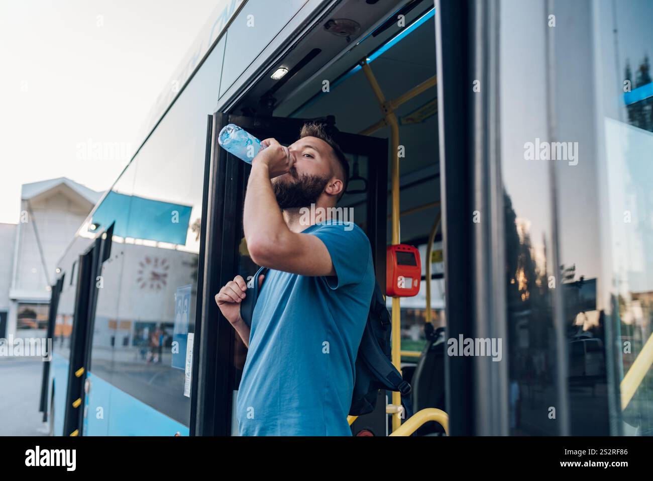 Giovane uomo allegro che beve una bottiglia d'acqua mentre scendi dall'autobus. Bell'uomo che prende l'autobus per andare al lavoro. Concetto di trasporto pubblico urbano. Copia spac Foto Stock