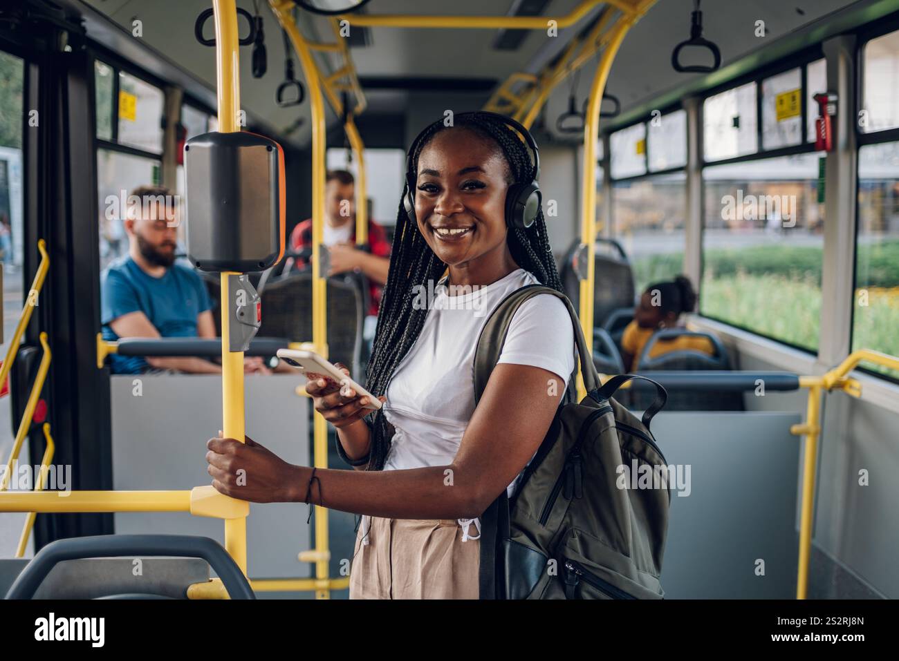 Giovane adorabile donna afroamericana gioiosa con trecce in piedi sull'autobus e che usa il telefono e guarda la telecamera. Trasporti pubblici e Foto Stock