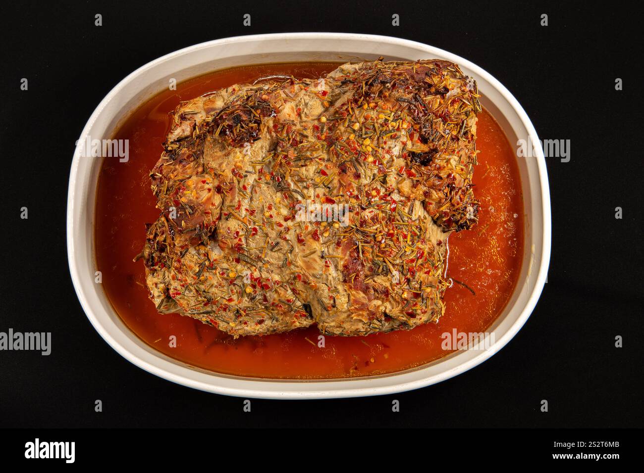 Una ciotola bianca ripiena di maiale arrosto imbevuta nei propri succhi salati, ricoperta di peperoncino rosso e rosmarino, su sfondo nero. Foto Stock