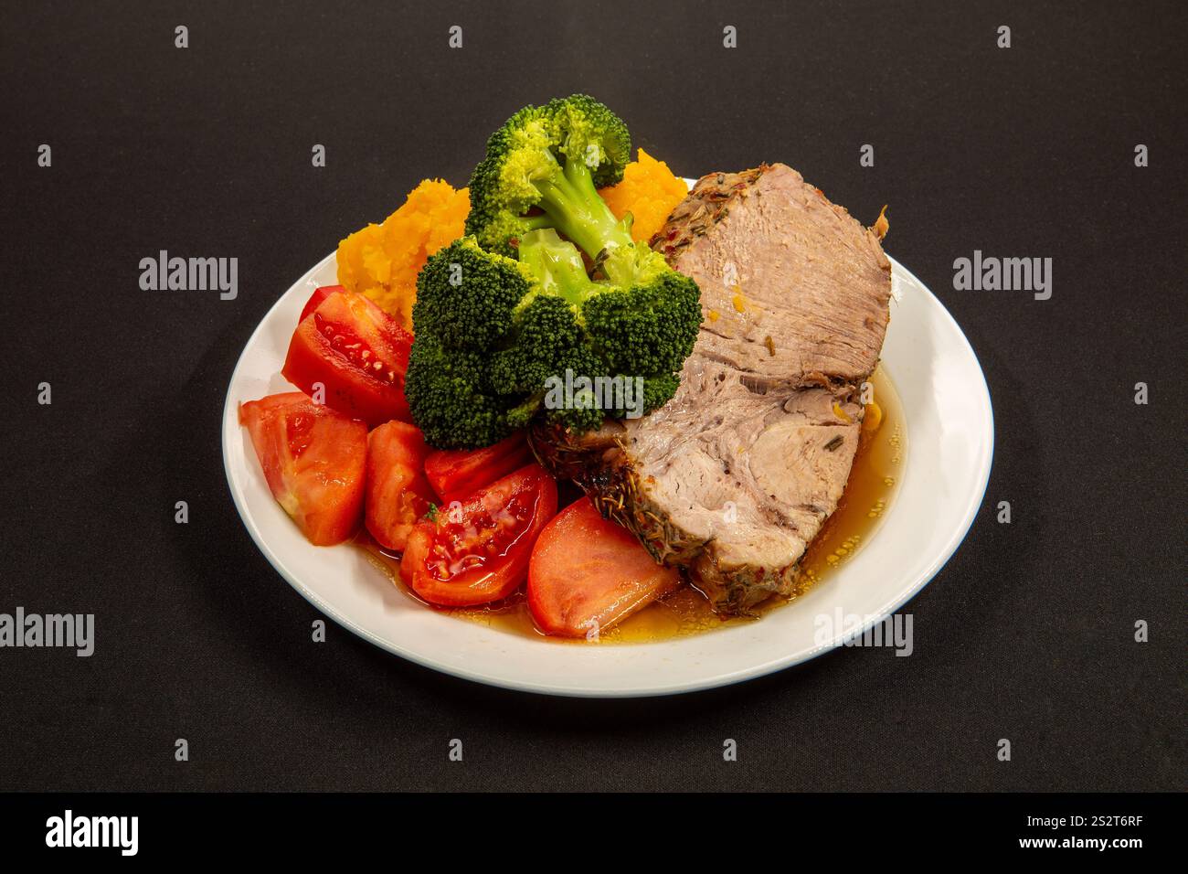 Spessa e succosa fetta di maiale arrosto, accompagnata da pomodori maturi e broccoli verdi, disposti su un semplice piatto bianco. Vista laterale. Sfondo nero. RIS Foto Stock
