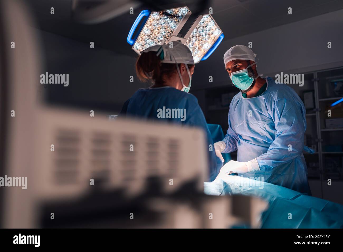 Sotto le luminose luci chirurgiche in sala operatoria, i chirurghi professionisti che indossano abiti e cappucci blu eseguono un'operazione con messa a fuoco e preci intensi Foto Stock