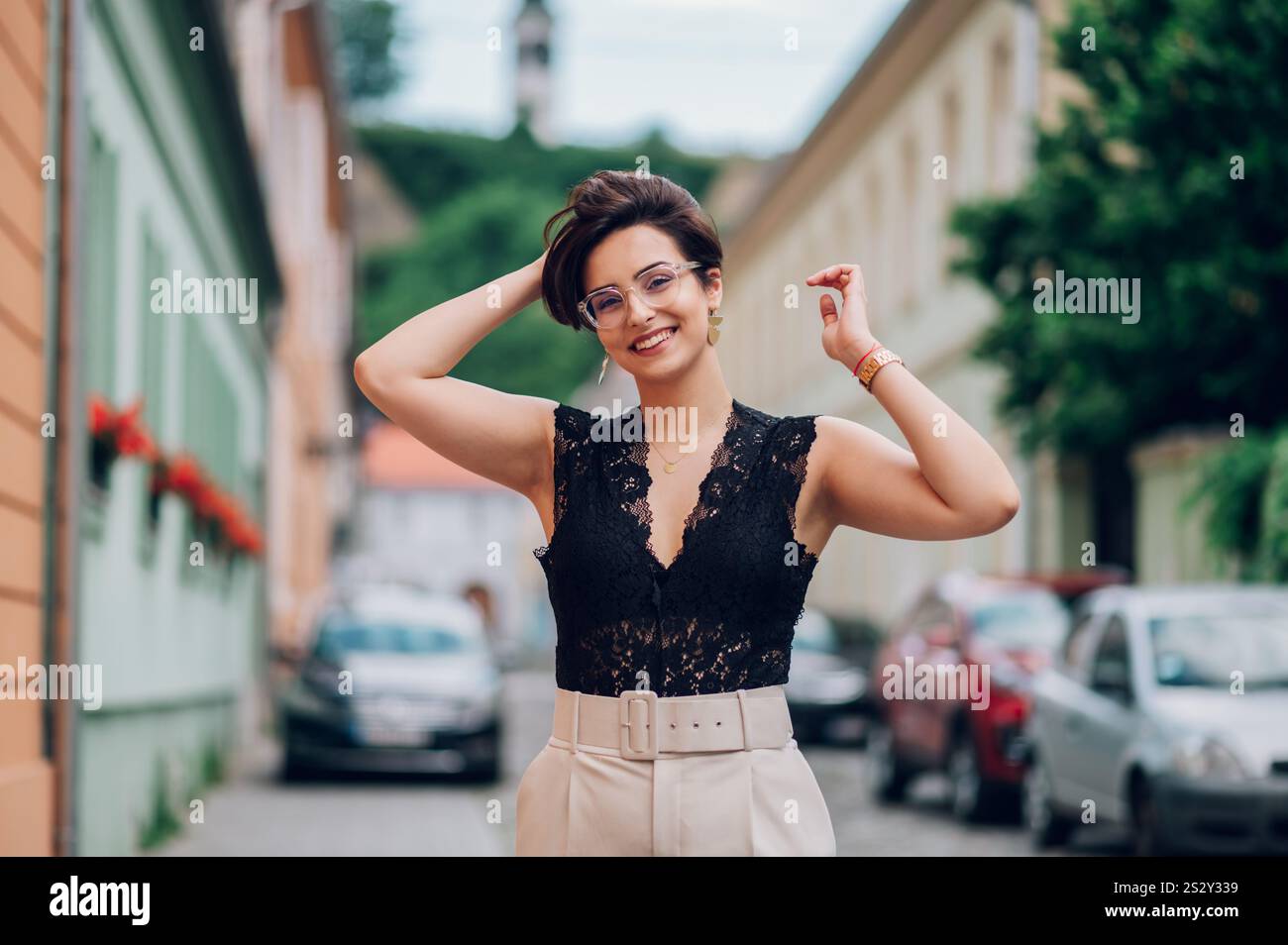 Bella giovane donna in abiti eleganti che posa all'aperto toccando i capelli con le mani. Ritratto di una donna attraente di buon umore che cammina Foto Stock