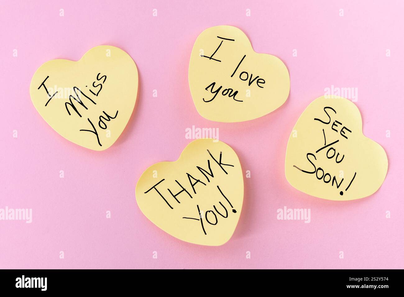 Quattro post-it gialli a forma di cuore con messaggi scritti a mano: "Ti amo", "mi manchi", "Grazie" e "a presto", disposti su un pastello Foto Stock