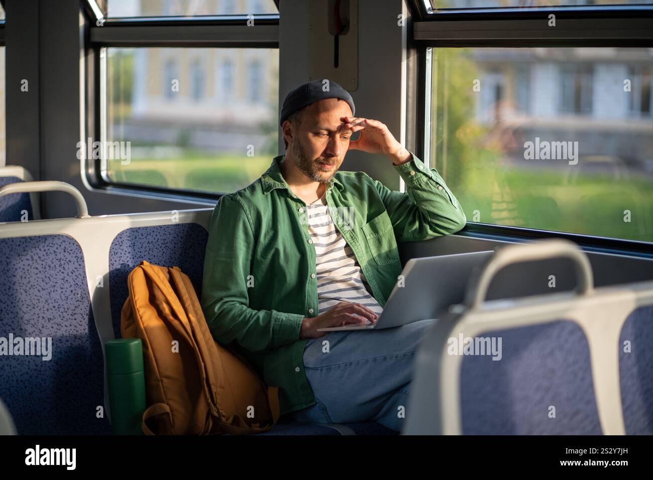 L'uomo lavora con un notebook in treno, stanco del viaggio, occhi chiusi per proteggersi dal sole. Portabilità di viaggio Foto Stock