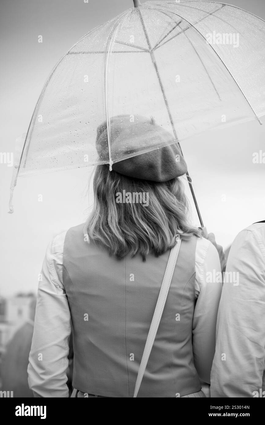 Vista posteriore monocromatica di una donna che indossa un gilet e un berretto e tiene in mano un ombrello trasparente al Goodwood Revival storico meeting Motorsport, Sussex, Regno Unito. Foto Stock