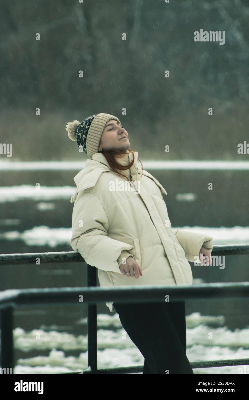 Una giovane donna in abbigliamento invernale si appoggia contro una ringhiera con gli occhi chiusi e un sorriso gentile. Foto Stock