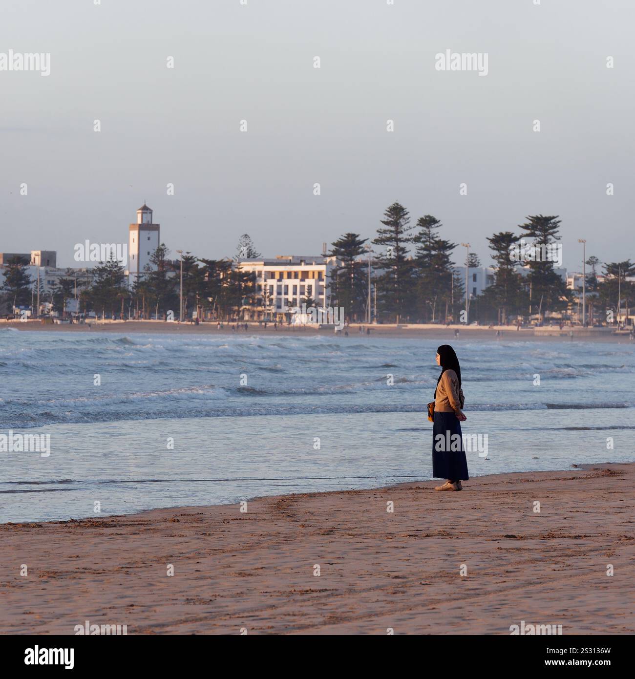 Una donna musulmana si erge su una spiaggia che guarda al mare mentre le onde si infrangono con la Medina dietro la città di Essaouira, in Marocco. Gennaio 2025 Foto Stock