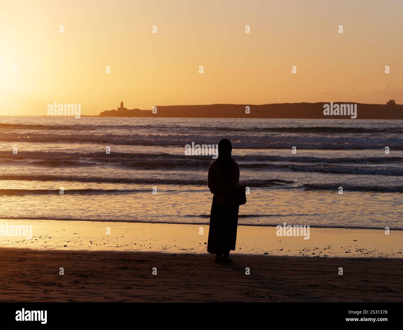 La donna musulmana si erge su una spiaggia che guarda al mare con l'isola all'orizzonte mentre le onde si infrangono. Essaouira, Marocco. Gennaio 2025 Foto Stock