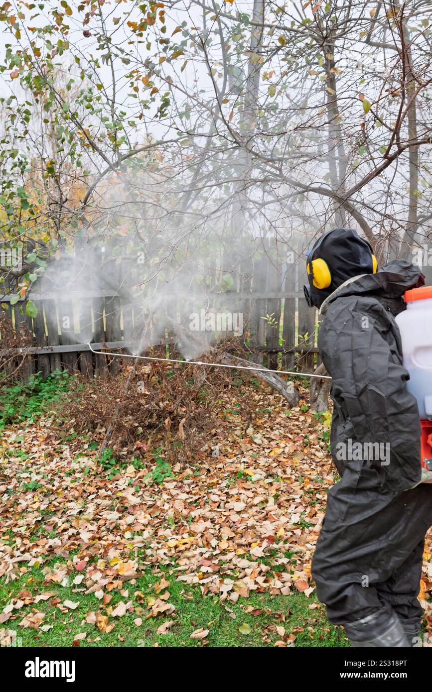 l'uomo in maschera e tuta spruzza il giardino con pesticidi e insetticidi contro i parassiti in autunno. Prevenzione di malattie in giardino. Trattamento stagionale Foto Stock