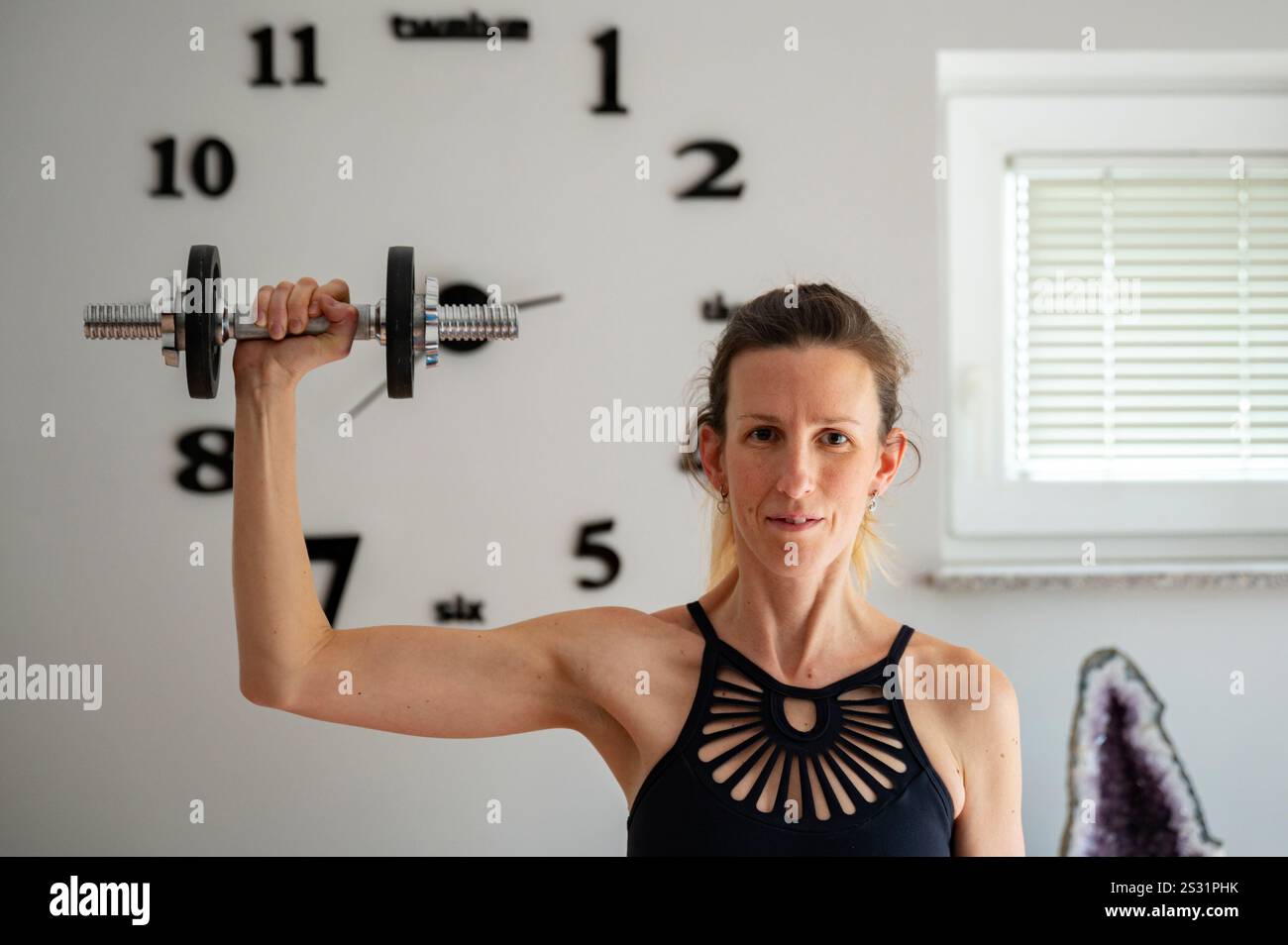 Una donna che solleva un manubrio in una stanza ben illuminata con un orologio decorativo e un cristallo di ametista sullo sfondo, incarnando forza e determinazione. Foto Stock