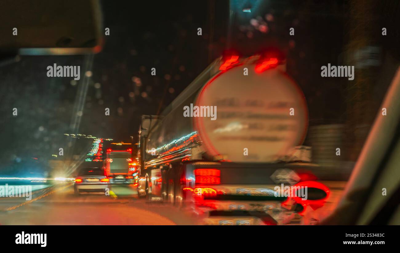 Vista sfocata da un'auto che guida dietro un camion su un'autostrada piovosa di notte. Concetto di percorrenza a lunga distanza e condizioni stradali notturne Foto Stock