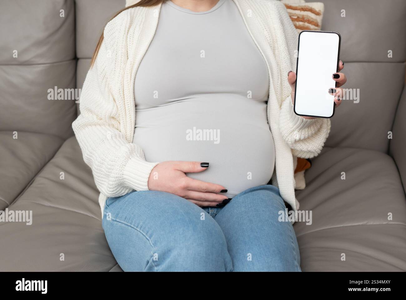 Primo piano di una donna incinta che tiene e mostra il telefono cellulare con display vuoto a casa. Immagine mockup con spazio di copia per app o interfaccia Foto Stock