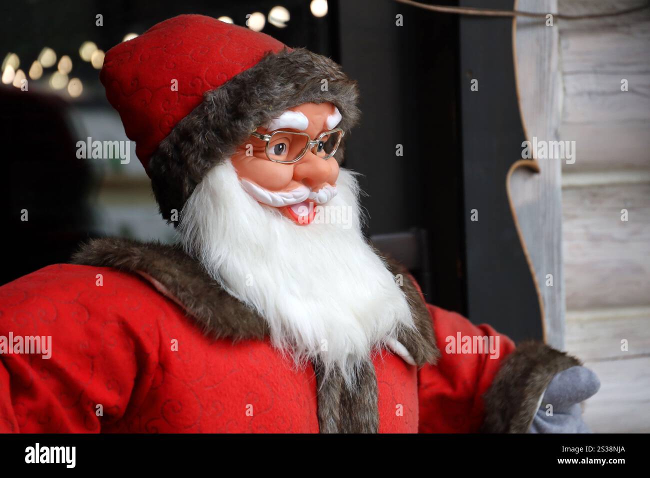 Grande figura di plastica di Babbo natale con abiti classici e cappello di babbo natale vicino alla casa di legno. Grande personaggio di plastica di Babbo natale con abiti classici di babbo natale Foto Stock