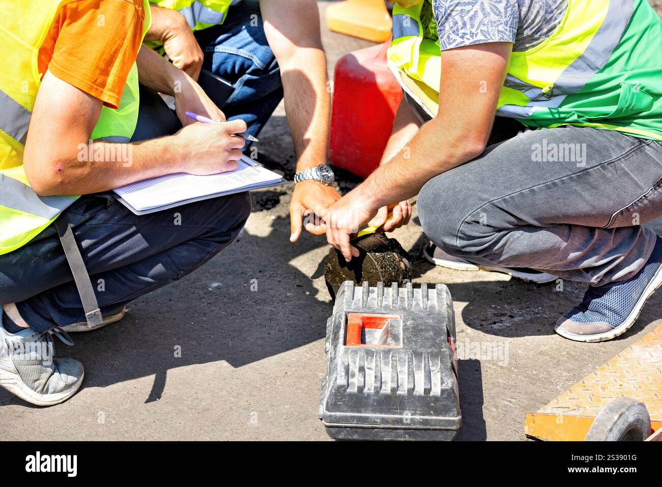 Un gruppo di operai edili in giubbotti fluorescenti si raduna intorno a una zona di strada, esaminando campioni. Foto Stock