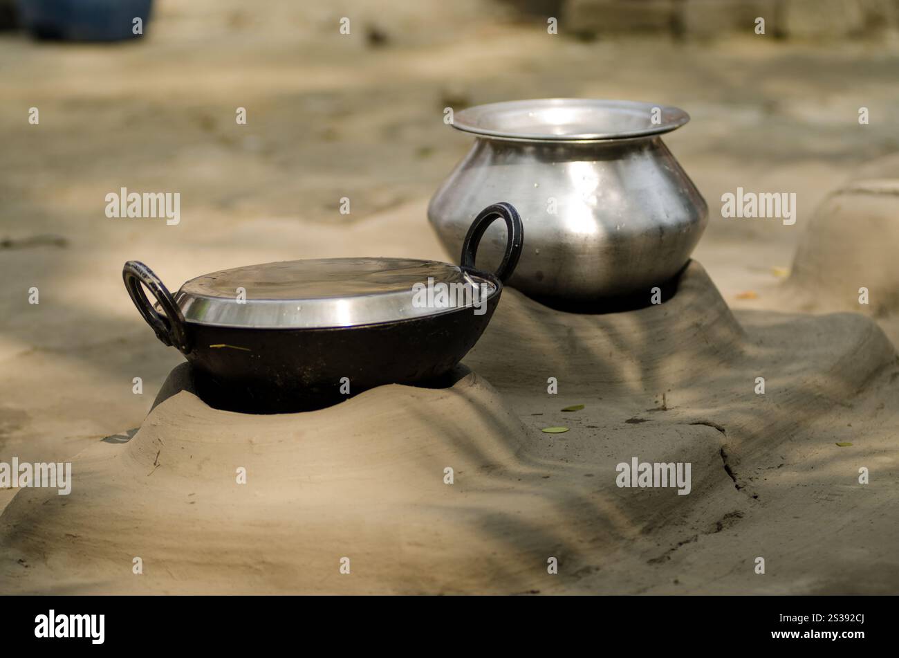Due utensili da cucina, una pentola nera e una ciotola argentata, poggiano su una superficie fangosa illuminata dalla luce soffusa del sole in un'area rurale all'aperto. Foto Stock
