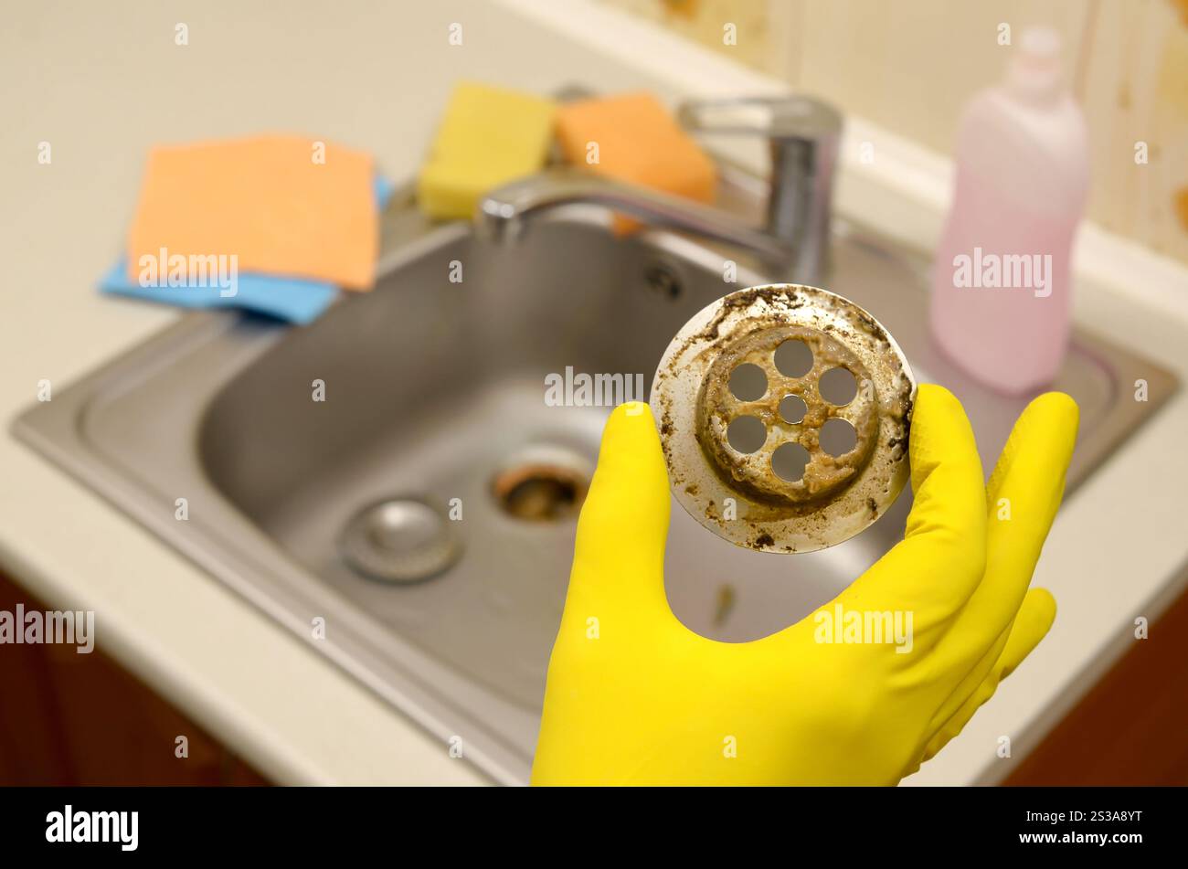 Il detergente nei guanti in gomma mostra i rifiuti nella protezione per peluche di un lavello da cucina. Filtro di scarico del lavandino in acciaio sporco. I rifiuti intrappolano la griglia dei fori di drenaggio. Foto Stock