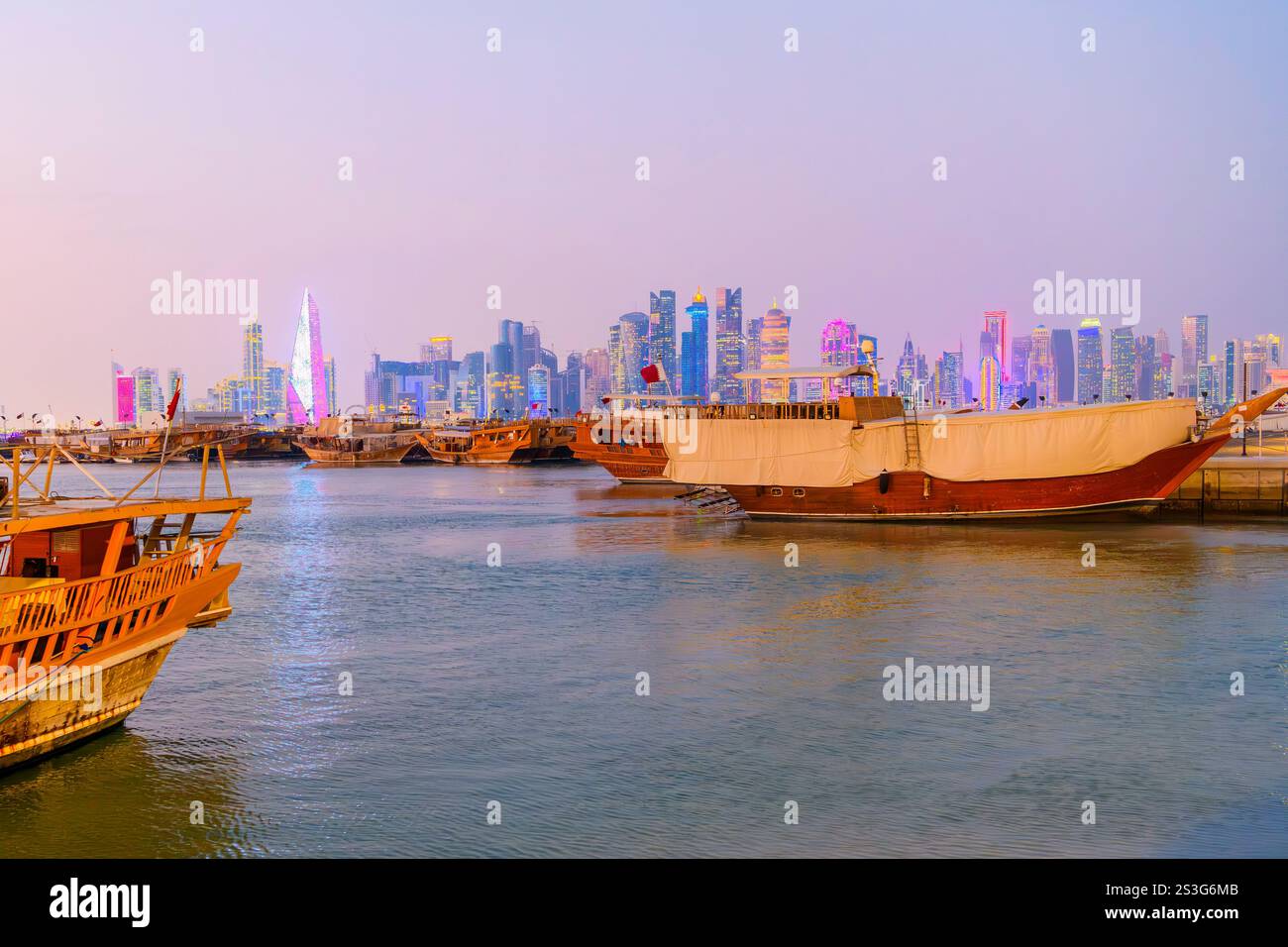 Barche tradizionali in dhow lungo la Corniche sul lungomare con lo skyline illuminato in lontananza all'ora blu crepuscolo, a Doha in Qatar. Foto Stock